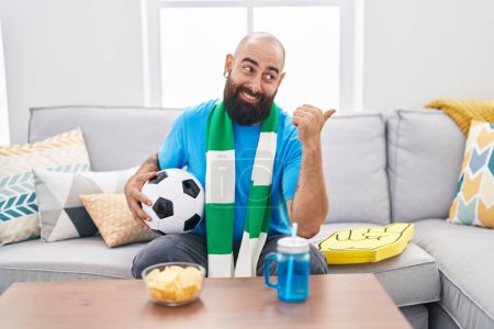 Photo for Young hispanic man with beard and tattoos football hooligan holding ball supporting team pointing thumb up to the side smiling happy with open mouth - Royalty Free Image