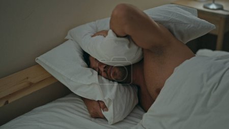 Téléchargez les photos : Jeune homme hispanique couvrant les oreilles pour le bruit couché sur le lit dans la chambre - en image libre de droit