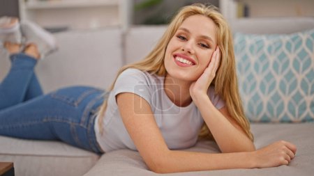 Foto de Mujer rubia joven sonriendo acostada en el sofá en casa - Imagen libre de derechos
