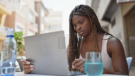 Téléchargez les photos : Femme afro-américaine utilisant un ordinateur portable assis sur la table à la terrasse du café - en image libre de droit