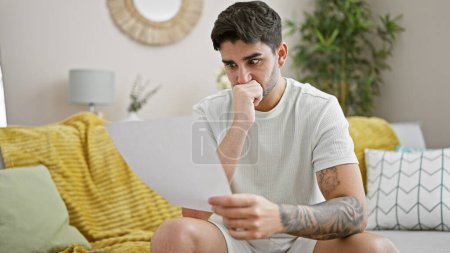 Téléchargez les photos : Jeune homme hispanique lisant le document assis sur le canapé à la maison - en image libre de droit