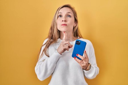 Photo for Young blonde woman using smartphone typing message thinking concentrated about doubt with finger on chin and looking up wondering - Royalty Free Image