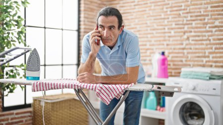 Foto de Hombre de mediana edad hablando en el teléfono inteligente apoyado en la máquina de planchar en la sala de lavandería - Imagen libre de derechos