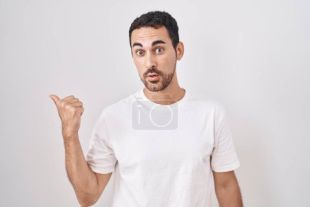 Photo for Handsome hispanic man standing over white background surprised pointing with hand finger to the side, open mouth amazed expression. - Royalty Free Image