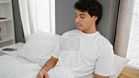 Foto de Hombre latino joven sentado en la cama pensando en el dormitorio - Imagen libre de derechos