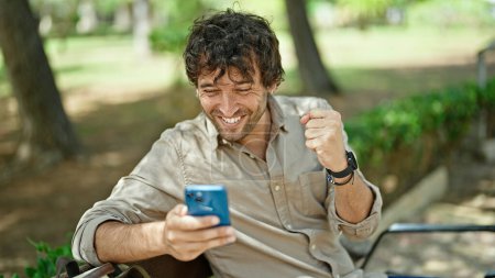 Foto de Joven hispano usando smartphone con gesto ganador sentado en el banco del parque - Imagen libre de derechos