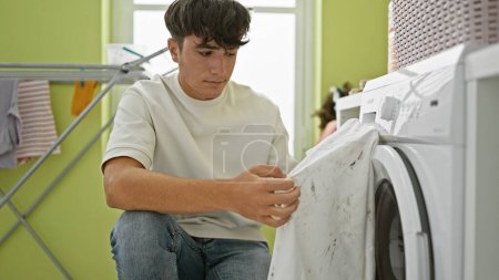 Photo for Hispanic young teenager seriously concentrating on tackling a dirty t-shirt stain, primed to clean it in the laundry room at home - Royalty Free Image