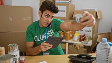 Téléchargez les photos : Jeune homme hispanique bénévole utilisant pavé tactile parler sur smartphone vérifier les produits au centre de charité - en image libre de droit