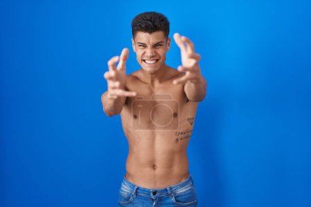 Foto de Joven hombre hispano de pie sin camisa sobre fondo azul gritando frustrado de rabia, manos tratando de estrangular, gritando loco - Imagen libre de derechos