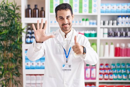 Foto de Hombre hispano guapo trabajando en farmacia mostrando y señalando con los dedos el número seis mientras sonríe confiado y feliz. - Imagen libre de derechos