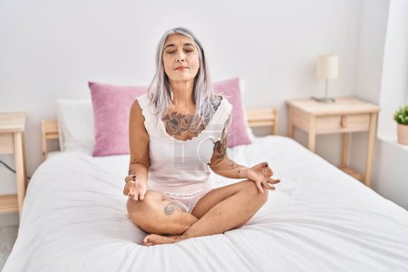 Téléchargez les photos : Moyen âge femme aux cheveux gris faisant de l'exercice de yoga assis sur le lit dans la chambre - en image libre de droit
