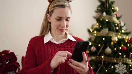 Photo for Young blonde woman using smartphone standing by christmas tree at home - Royalty Free Image