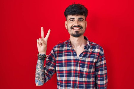 Téléchargez les photos : Jeune homme hispanique avec la barbe debout sur fond rouge montrant et pointant vers le haut avec les doigts numéro deux tout en souriant confiant et heureux. - en image libre de droit
