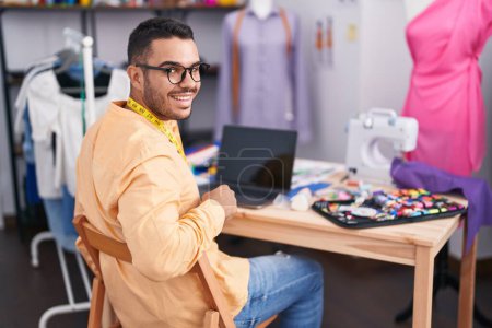 Téléchargez les photos : Jeune homme hispanique tailleur souriant confiant en utilisant un ordinateur portable au magasin de tailleur - en image libre de droit