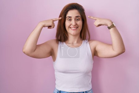 Photo for Brunette woman standing over pink background smiling pointing to head with both hands finger, great idea or thought, good memory - Royalty Free Image
