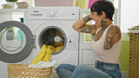 Photo for Hispanic woman with amputee arm washing clothes sitting on floor stressed at laundry room - Royalty Free Image