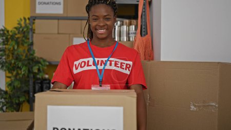 Téléchargez les photos : Femme afro-américaine bénévole souriant confiant tenant paquet de dons au bureau - en image libre de droit