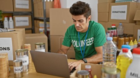 Téléchargez les photos : Jeune homme hispanique bénévole utilisant un ordinateur portable avec un visage sérieux au centre de charité - en image libre de droit