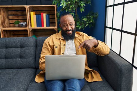 Téléchargez les photos : Afro-Américain utilisant ordinateur portable à la maison assis sur le canapé souriant heureux pointant avec la main et le doigt - en image libre de droit