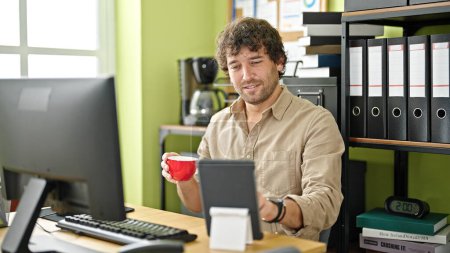 Foto de Joven trabajador de negocios hispano usando touchpad bebiendo café en la oficina - Imagen libre de derechos