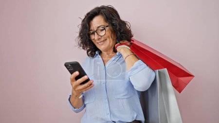 Foto de Mujer hispana de mediana edad usando teléfono inteligente sosteniendo bolsas de compras sobre un fondo rosa aislado - Imagen libre de derechos