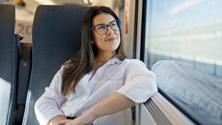 Foto de Joven hermosa mujer hispana sonriendo feliz mirando a través de la ventana dentro de vagón de tren - Imagen libre de derechos