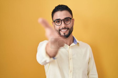 Foto de Joven hispano vistiendo ropa de negocios y gafas sonriente alegre ofreciendo mano de palma dando asistencia y aceptación. - Imagen libre de derechos