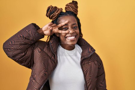 Photo for African woman with braided hair standing over yellow background doing peace symbol with fingers over face, smiling cheerful showing victory - Royalty Free Image