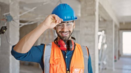 Joven constructor hispano sonriendo confiado usando el saludo del hardhat en el sitio de construcción