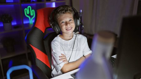 Photo for Cute blond boy streamer, engrossed in a digital game, sitting cross-armed and smiling in a dark gaming room lit up by futuristic virtual entertainment technology. - Royalty Free Image
