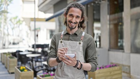 Téléchargez les photos : Jeune homme hispanique serveur souriant confiant prendre des notes à la terrasse du café - en image libre de droit