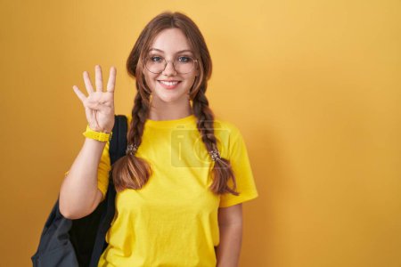 Foto de Joven mujer caucásica usando mochila de estudiante sobre fondo amarillo mostrando y apuntando hacia arriba con los dedos número cuatro mientras sonríe confiado y feliz. - Imagen libre de derechos