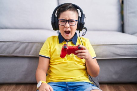 Photo for Young hispanic kid playing video game holding controller wearing headphones angry and mad screaming frustrated and furious, shouting with anger. rage and aggressive concept. - Royalty Free Image