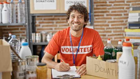 Photo for Young hispanic man volunteer writing on clipboard smiling at charity center - Royalty Free Image