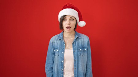 Foto de Mujer joven caucásica expresión sorpresa usando sombrero de Navidad sobre fondo rojo aislado - Imagen libre de derechos