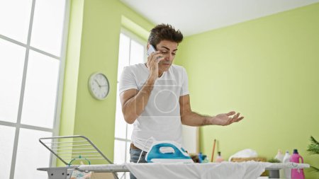Foto de Joven hombre hispano hablando en smartphone planchando ropa en la lavandería - Imagen libre de derechos