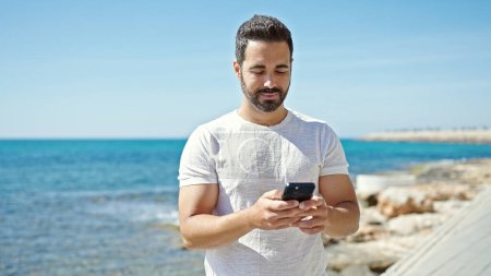 Foto de Joven hombre hispano sonriendo confiado usando smartphone en la playa - Imagen libre de derechos