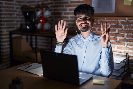 Téléchargez les photos : Jeune homme hispanique avec la barbe travaillant au bureau la nuit montrant et pointant vers le haut avec les doigts numéro huit tout en souriant confiant et heureux. - en image libre de droit