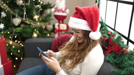 Foto de Joven mujer hispana hermosa usando teléfono inteligente y tarjeta de crédito sentado en el sofá por el árbol de Navidad en casa - Imagen libre de derechos