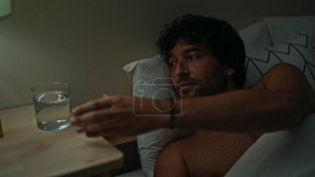 Téléchargez les photos : Jeune homme hispanique tenant un verre d'eau allongé sur le lit torse nu dans la chambre - en image libre de droit