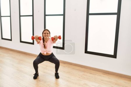 Téléchargez les photos : Jeune belle femme hispanique souriant confiant en utilisant haltères formation au centre sportif - en image libre de droit