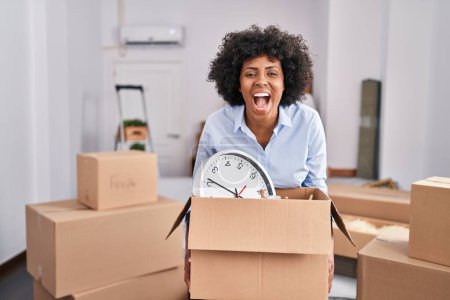 Téléchargez les photos : Femme noire avec les cheveux bouclés se déplaçant vers une nouvelle maison tenant boîte en carton souriant et riant dur à haute voix parce que drôle blague folle. - en image libre de droit