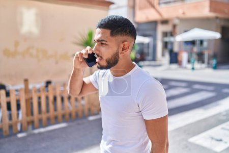 Téléchargez les photos : Jeune homme latin parlant sur smartphone avec une expression sérieuse à la rue - en image libre de droit