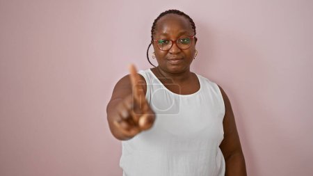 Téléchargez les photos : Femme afro-américaine résiliente, plus la taille, debout dans un style décontracté, disant "non" avec une expression sérieuse et lever le doigt sur fond rose froid isolé. - en image libre de droit