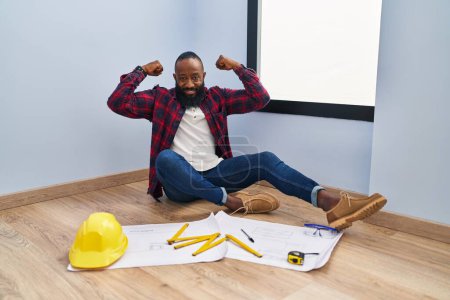 Photo for African american man sitting on the floor at new home looking at blueprints showing arms muscles smiling proud. fitness concept. - Royalty Free Image