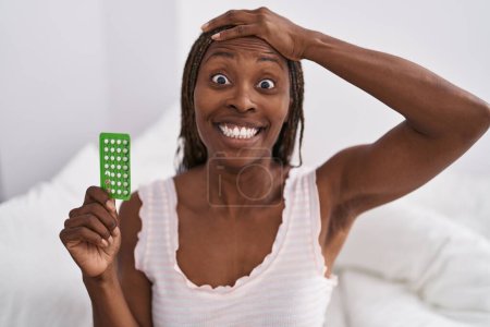 Foto de Mujer afroamericana sosteniendo píldoras anticonceptivas estresada y frustrada con la mano en la cabeza, cara sorprendida y enojada - Imagen libre de derechos