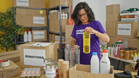 Photo for Middle age hispanic woman volunteer putting products on cardboard box to donate at charity center - Royalty Free Image