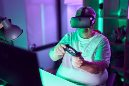 Téléchargez les photos : Homme aux cheveux gris moyen âge streamer jouer à un jeu vidéo en utilisant des lunettes de réalité virtuelle et joystick à la salle de jeux - en image libre de droit