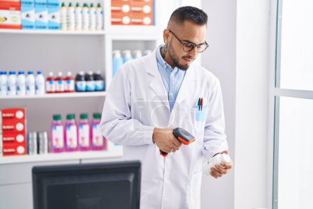 Téléchargez les photos : Jeune homme hispanique pharmacien numérisation pilules bouteille à la pharmacie - en image libre de droit