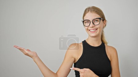 Téléchargez les photos : Jeune femme blonde souriante pointant vers le côté présentant sur fond blanc isolé - en image libre de droit